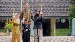 Keluarga yang bangga memegang piala wisuda dengan pakaian tradisional di luar sebuah bangunan.