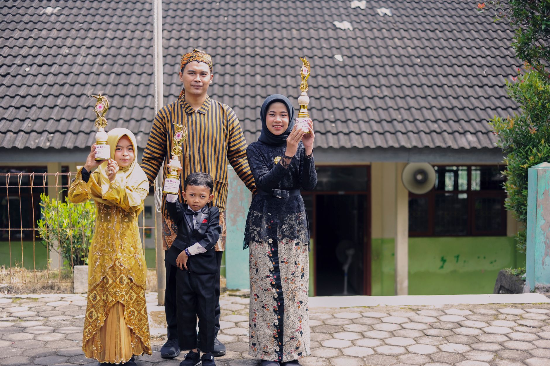 Keluarga yang bangga memegang piala wisuda dengan pakaian tradisional di luar sebuah bangunan.