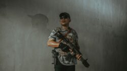Seorang tentara mengenakan perlengkapan taktis dan kacamata hitam memegang senapan serbu di dalam ruangan.