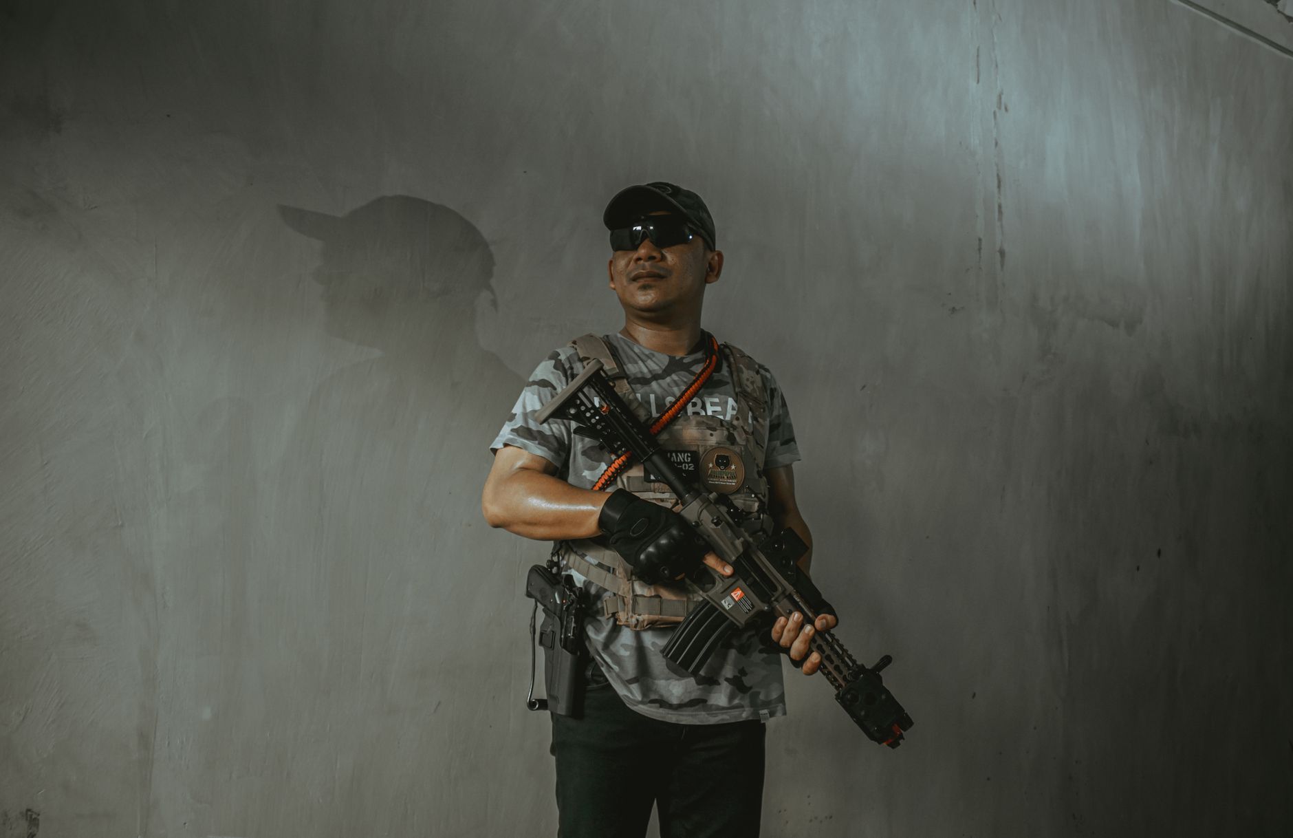 Seorang tentara mengenakan perlengkapan taktis dan kacamata hitam memegang senapan serbu di dalam ruangan.