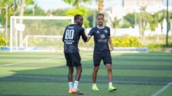 Dua pemain sepak bola saling menyapa saat latihan di lapangan luar ruangan yang cerah.