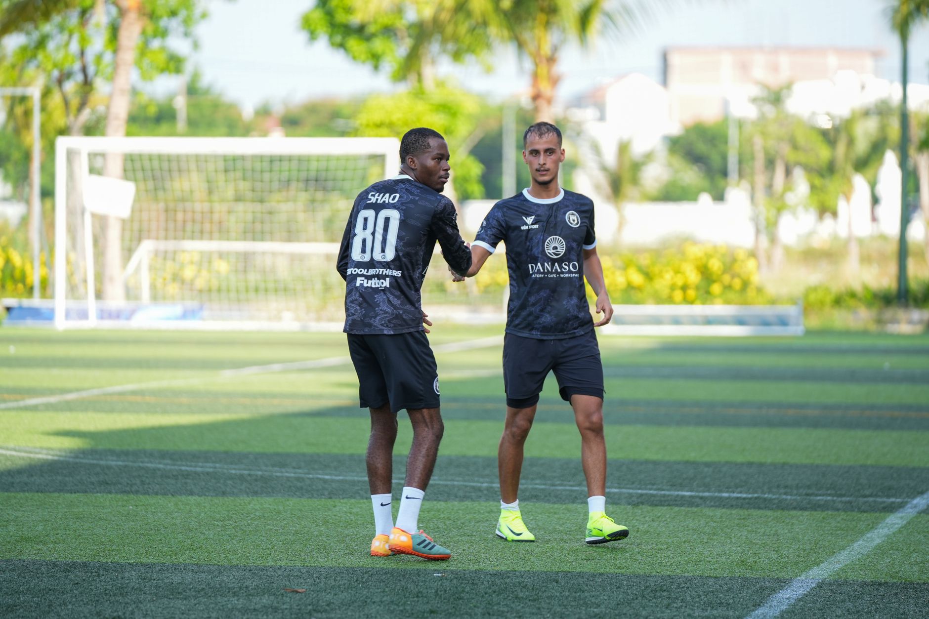 Dua pemain sepak bola saling menyapa saat latihan di lapangan luar ruangan yang cerah.