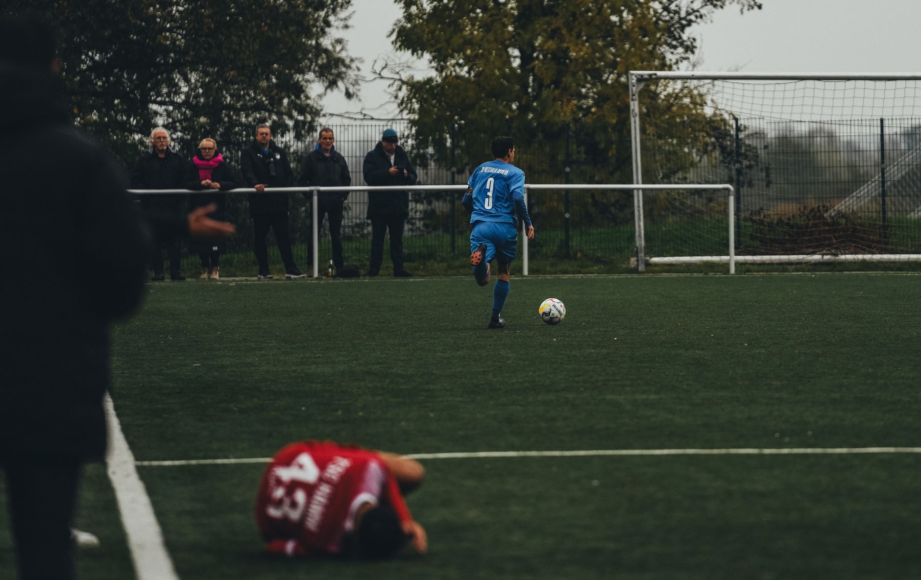 Seorang pemain sepak bola yang mengenakan biru berlari di lapangan sementara para penonton mengamati dengan seksama selama pertandingan yang intens.
