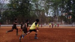 Sekelompok anak bermain sepak bola di lapangan tanah dengan pohon di latar belakang, menampilkan kerja sama tim dan semangat olahraga.