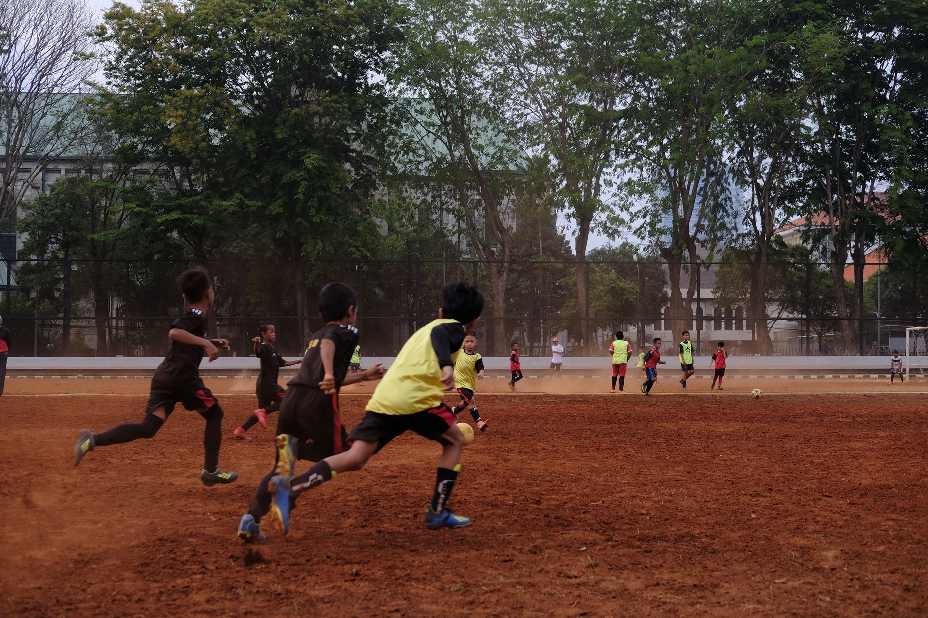 Sekelompok anak bermain sepak bola di lapangan tanah dengan pohon di latar belakang, menampilkan kerja sama tim dan semangat olahraga.