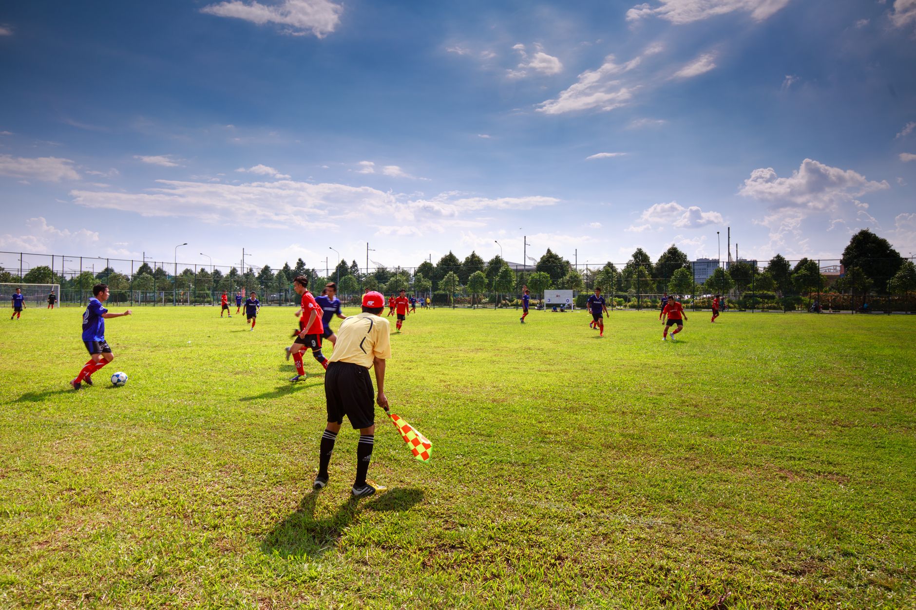 Pertandingan sepak bola remaja di lapangan terbuka dengan pemain, wasit, dan langit yang cerah.