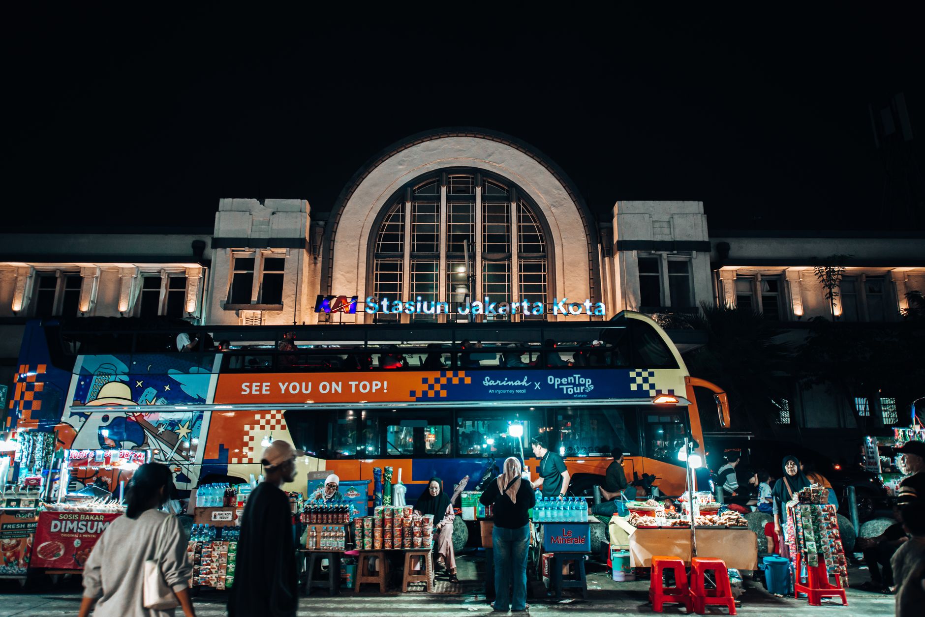 Suasana pasar malam yang ramai di Stasiun Jakarta Kota dengan bus tingkat yang mencolok dan para pedagang kaki lima.