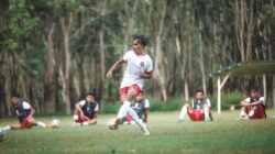 Pemain sepak bola yang dinamis dalam aksi selama sesi pelatihan luar ruangan dengan rekan tim di latar belakang.