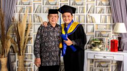Ayah dan anak merayakan kelulusan di lingkungan rumah yang nyaman dengan rak buku dan dekorasi.