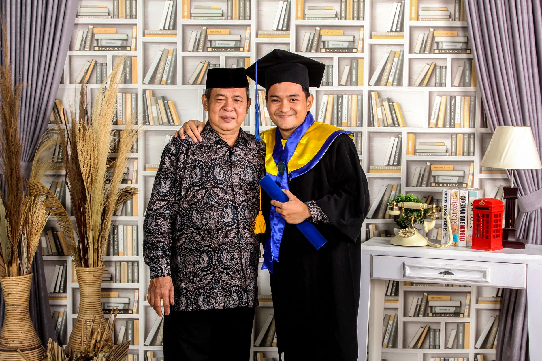 Ayah dan anak merayakan kelulusan di lingkungan rumah yang nyaman dengan rak buku dan dekorasi.