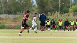 Pertandingan sepak bola amatir yang penuh aksi dengan para pemain yang bergerak dan seorang pelatih di pinggir lapangan.