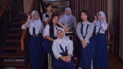 Tujuh gadis remaja berseragam sekolah berpose di dalam ruangan dengan suasana santai dan ramah.