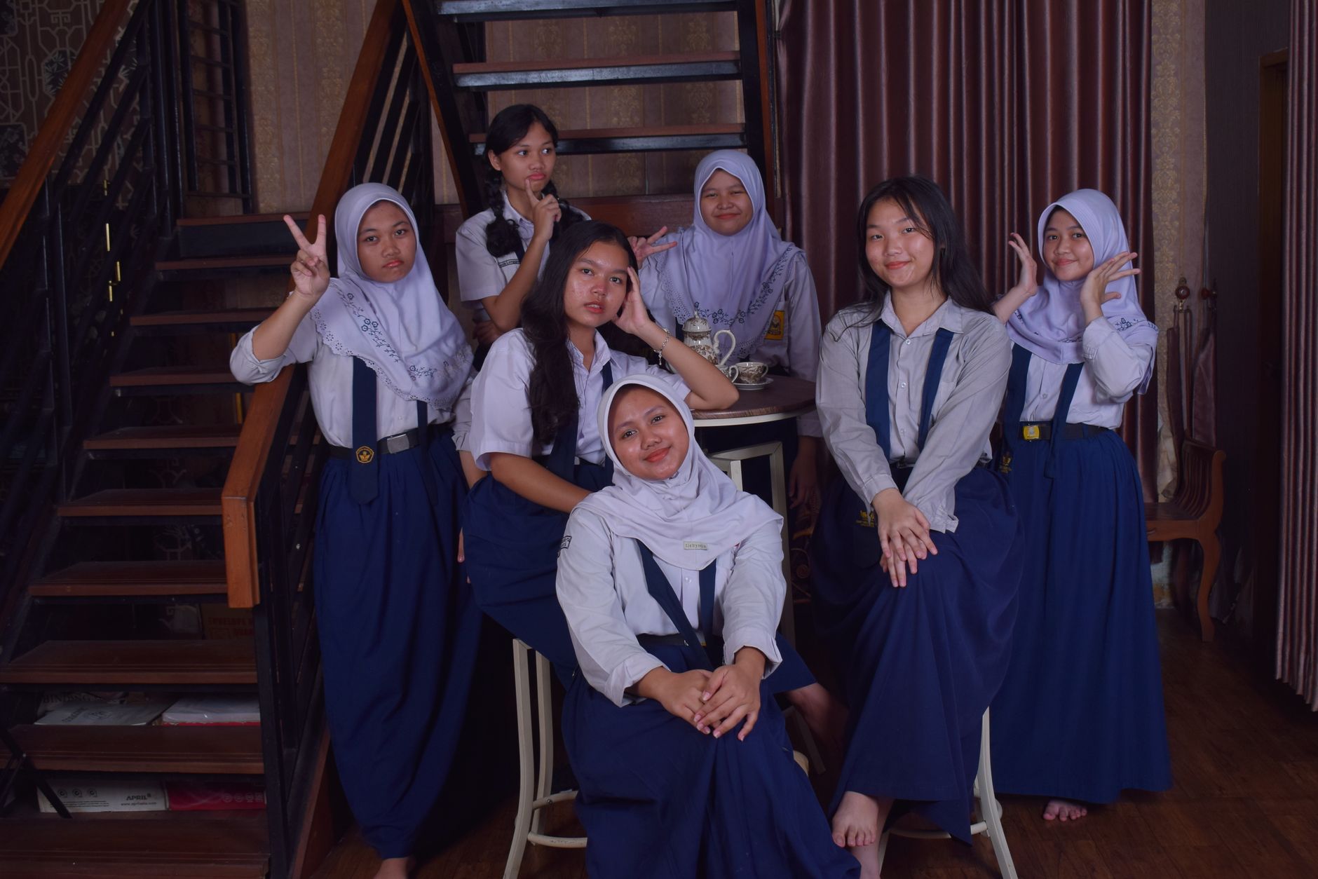 Tujuh gadis remaja berseragam sekolah berpose di dalam ruangan dengan suasana santai dan ramah.