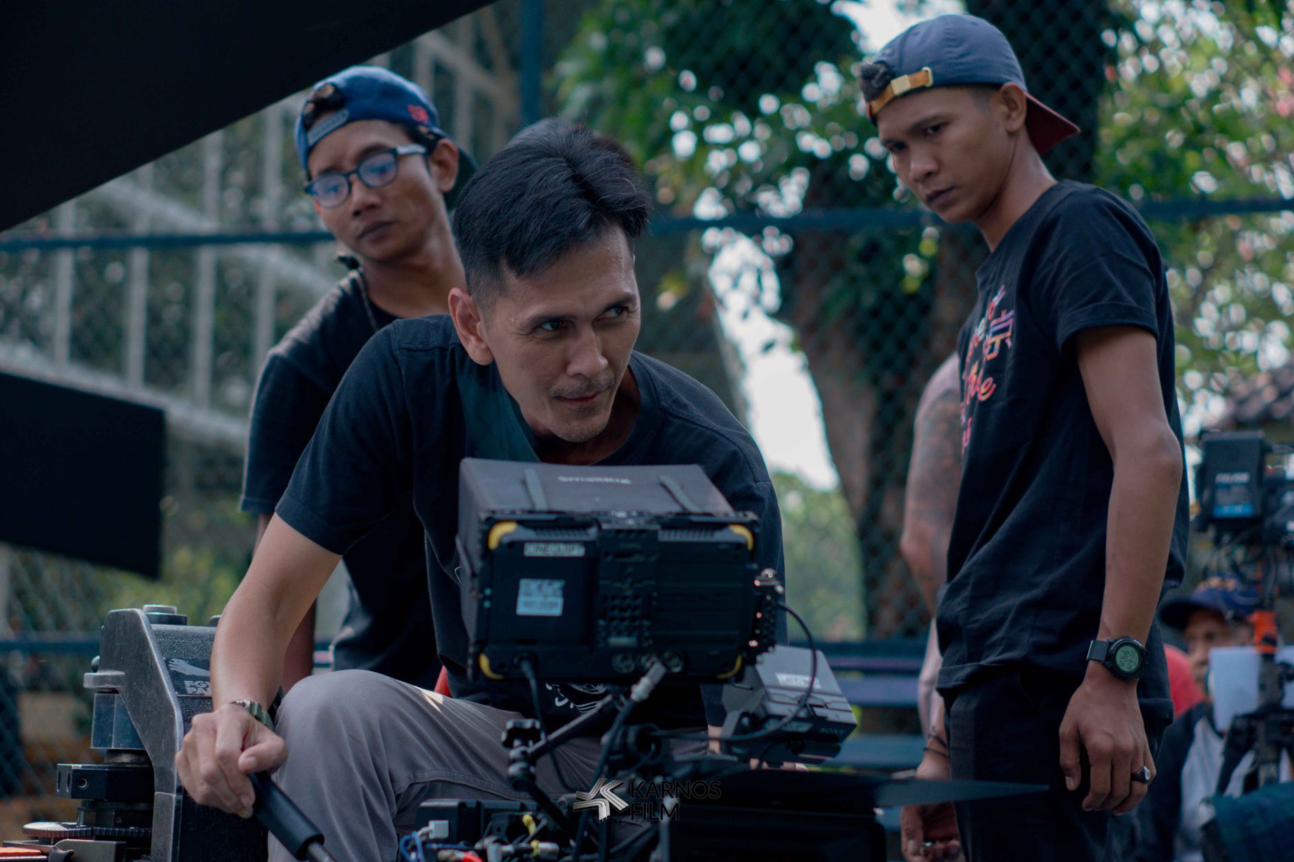 Sekelompok pembuat film yang meninjau rekaman di lokasi syuting luar ruangan, fokus pada perlengkapan kamera.
