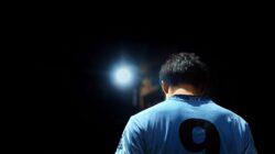 Seorang pemain sepak bola dengan jersey biru bernomor 9, diterangi oleh lampu sorot di malam hari.